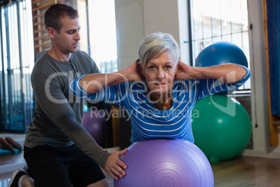 Physiotherapist assisting senior woman in performing exercise on fitness ball