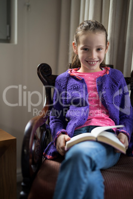 Smiling girl reading book on chair in living room