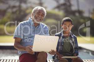 Smiling grandfather and grandson sitting together on bench with laptop and digital tablet