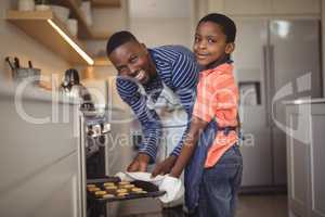 Father taking tray of fresh cookies out of oven with son in kitchen