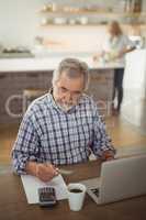 Senior man paying bills online on laptop in kitchen
