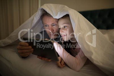 Father and daughter using digital tablet under blanket on bed