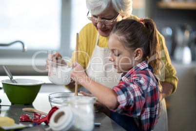 Grandmother adding water while granddaughter is mixing flour in a bowl