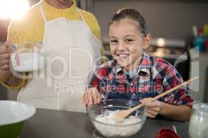 Grandmother adding water while granddaughter is mixing flour in a bowl