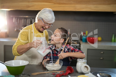 Grandmother adding water while granddaughter is mixing flour in a bowl