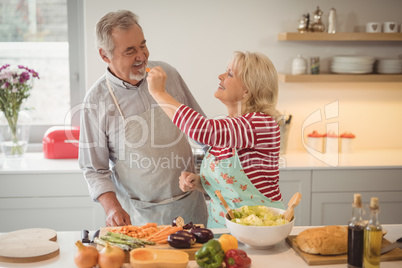 Senior woman feeding vegetable salad to man in kitchen