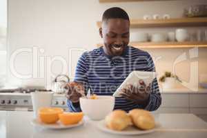 Smiling man using a digital tablet while having breakfast in kitchen