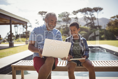 Smiling grandfather showing laptop to grandson near poolside