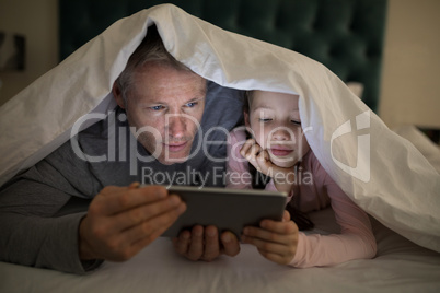 Father and daughter using digital tablet under blanket on bed