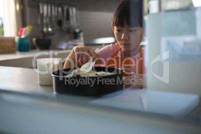 Girl spreading cream on cake seen through glass in kitchen