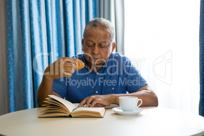Senior man eating croissant while reading book in nursing home