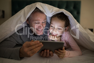 Father and daughter using digital tablet under blanket on bed