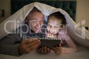 Father and daughter using digital tablet under blanket on bed