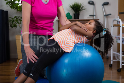 Female physiotherapist helping girl patient in performing exercise on fitness ball