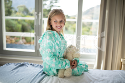 Smiling girl holding teddy bear on bed in bedroom