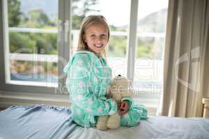Smiling girl holding teddy bear on bed in bedroom