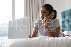 Woman talking on phone while using laptop on bed