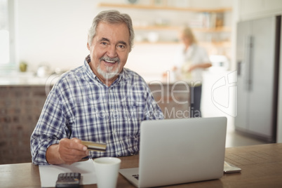 Smiling senior man paying bills online on laptop in kitchen