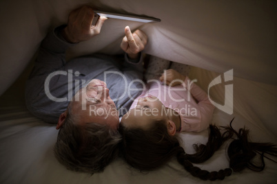 Father and daughter using digital tablet under blanket on bed