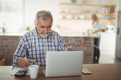 Senior man paying bills online on laptop in kitchen