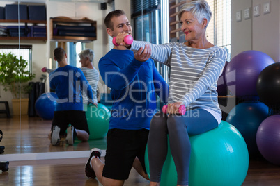 Physiotherapist assisting senior woman on exercise ball and dumbbells