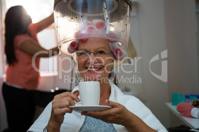Portrait of senior woman having drink while sitting under hair steamer