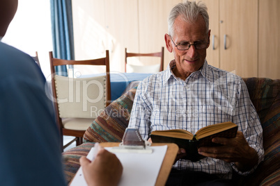 Doctor writing on paper while senior man reading book in nursing home