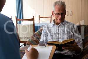 Doctor writing on paper while senior man reading book in nursing home