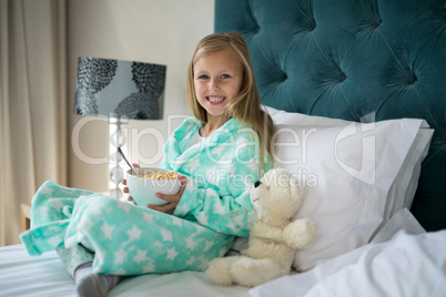 Smiling girl having breakfast with teady bear on bed