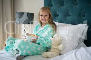 Smiling girl having breakfast with teady bear on bed