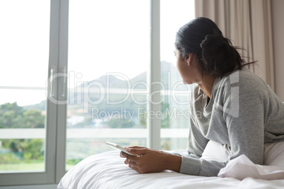 Woman looking away while holding digital tablet on bed