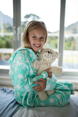 Smiling girl holding teddy bear on bed in bedroom