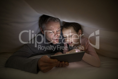 Father and daughter using digital tablet under blanket on bed