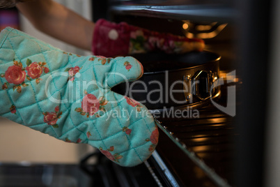 Cropped hands of girl wearing glove keeping container with cake in oven
