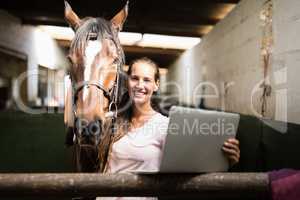 Portrait of female jockey holding laptop while standing by horse