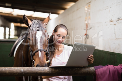 Smiling female jockey using laptop while standing by horse