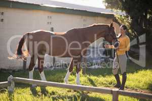 Full length of female jockey with horse standing on field at barn