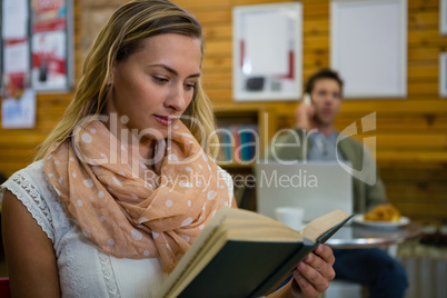 Woman reading book while man talking on phone in background