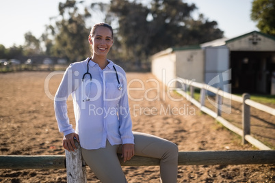Smiling female vet sitting on wooden fence at barn
