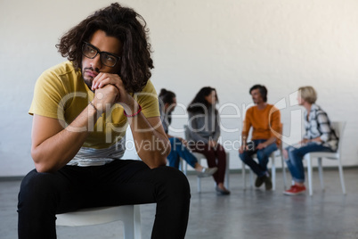 Man looking away while sitting on chair with friends discussing in background