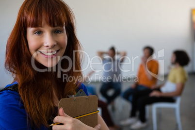 Portrait of female teacher holding clipboard with students talking in background