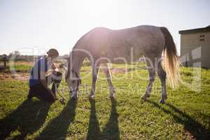 Female jockey looking at horse while kneeling on grassy field