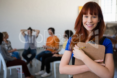 Portrait of happy teacher holding clipboard with students talking in background