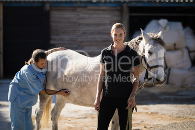 Female jockey standing by vet examining horse at barn