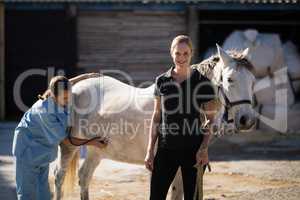 Female jockey standing by vet examining horse at barn