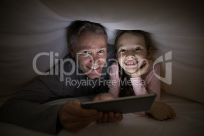 Smiling father and daughter with digital tablet under blanket on bed