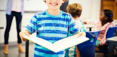 Portrait of schoolboy standing with book in classroom