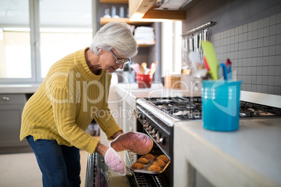 Senior woman taking tray of fresh cookies out of oven in kitchen