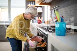 Senior woman taking tray of fresh cookies out of oven in kitchen