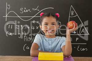 Happy student girl at table holding an apple against grey blackboard with education and school graph